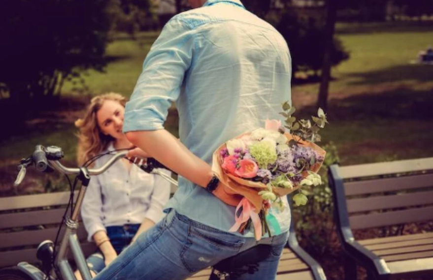 Man on e-bike hiding flower bouquet behind back, woman sitting on park bench, Pogo Cycles